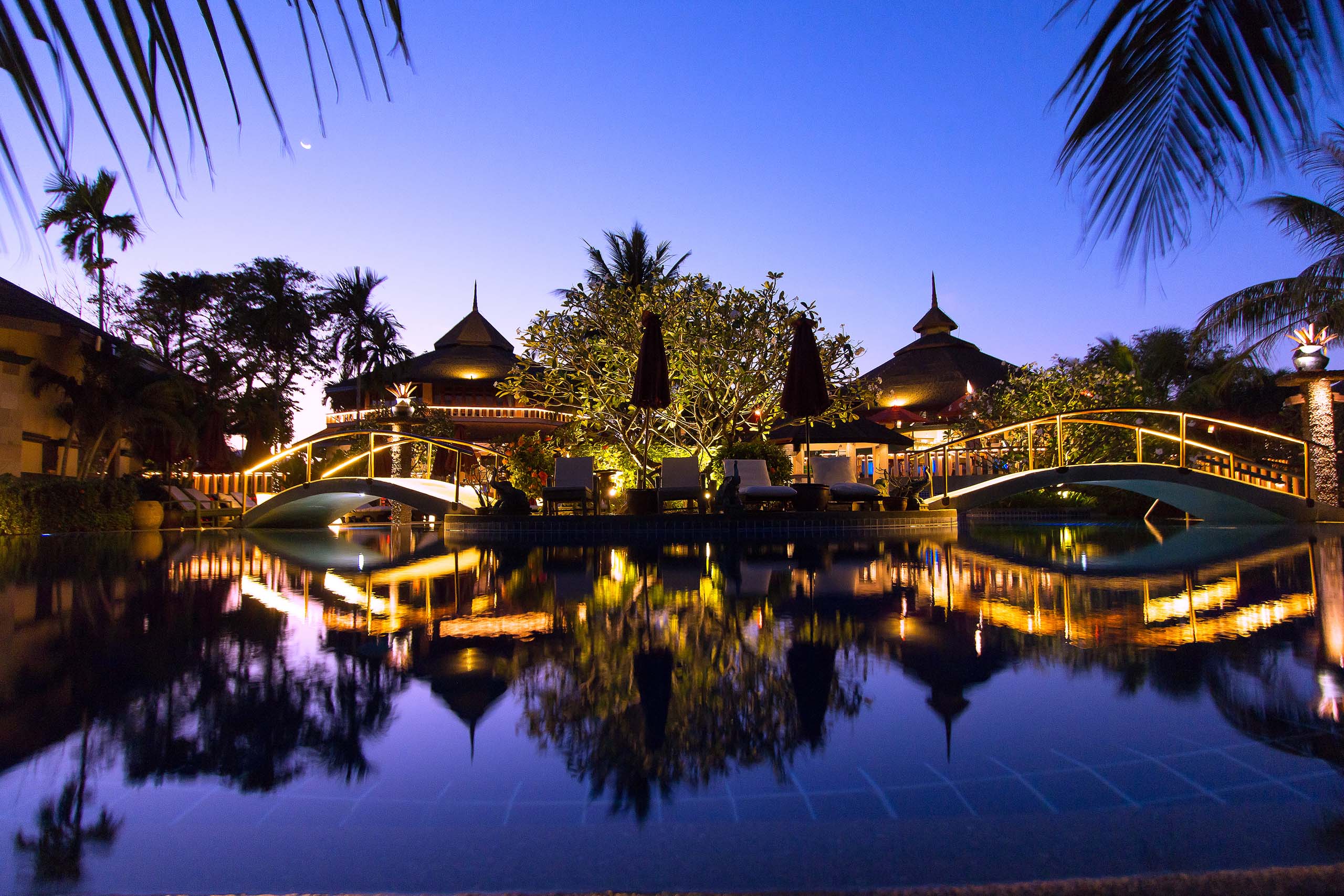 Resort pavilions and pool at dusk, interior lights reflected in the water